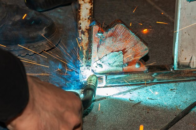 Una persona esperta esegue lavori con una saldatrice che fissa parti metalliche rimuovendo fumo blu e scintille gialle e fulmini nell'officina di produzione industriale