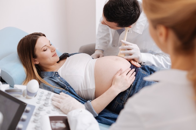 Experienced qualified skilled physicians working in the clinic while using medical equipment for ultrasound monitoring of pregnant belly and checking echo waves