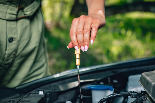 Experienced woman is checking an oil in the car via an oil dipstick against the background of nature, close up