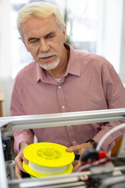 Foto esperto nel settore. piacevole uomo anziano che tiene una bobina di filamento per stampante 3d e controlla le prestazioni di una stampante 3d