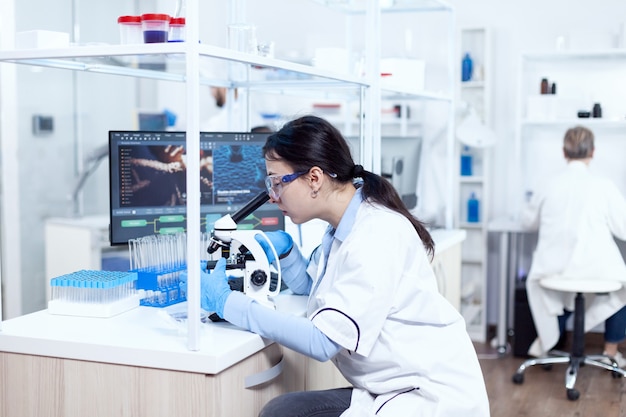 Expert in genetics doing research using microscope to analise chemical from human adn. chemist wearing lab coat using modern technology during scientific experiment in sterile environment.