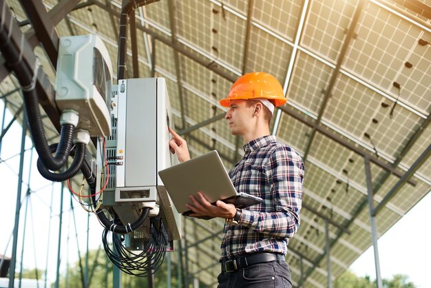 Expert is inspecting quality of solar panels Man in hardhat standing with laptop near electrical equipment