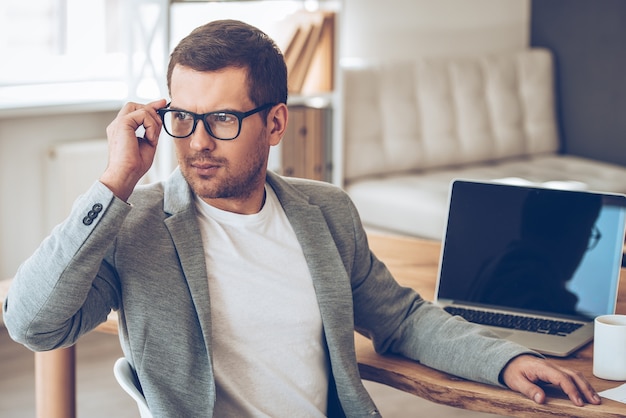 Expert look. Handsome young man adjusting his glasses and looking away while sitting at his working place