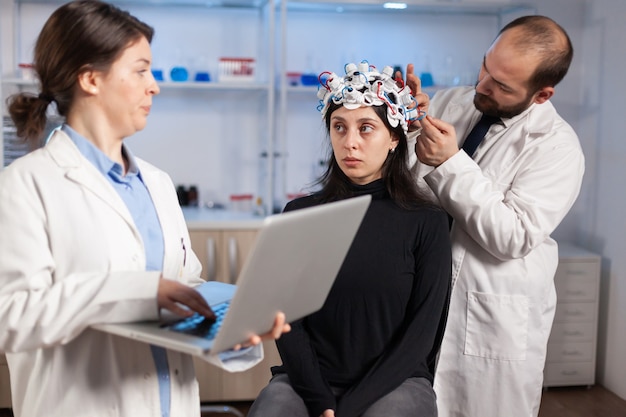 Photo expert medic in neuroscience developing brain experiment holding laptop explaning to woman side effects of nervous system treamtment. team of scientists working in neurological research laboratory.