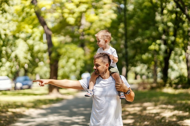 Exploring nature, father and son are playing in the park Father carries his son on his shoulders and they walk through the park one summer sunny day Dressed in the same clothes, dad point to something