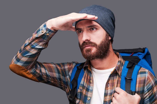 Photo exploring new places. handsome young bearded man carrying backpack and looking at view while holding hand upon his eyes and standing against grey background