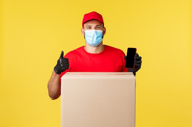 Photo express delivery during pandemic, covid-19, safe shipping, online shopping concept. courier in red uniform and medical mask, showing application to track orders, stand near box, hold smartphone