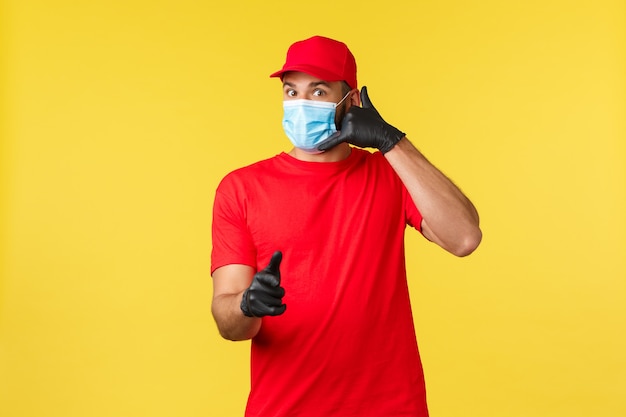 Consegna espressa durante la pandemia, covid-19, spedizione sicura, concetto di shopping online. corriere bello in uniforme rossa, maschera medica e guanti, indicando il cliente e facendo il segnale di chiamata