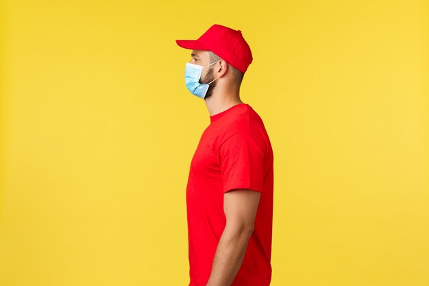 Express delivery during pandemic, covid-19, safe shipping, online shopping concept. profile of cheerful courier in red uniform and medical mask looking left, work with clients, taking package