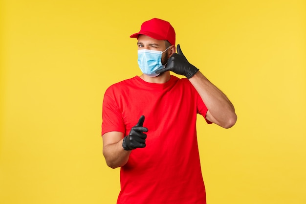 Express delivery during pandemic, covid-19, safe shipping, online shopping concept. Friendly courier in red uniform, medical mask and gloves, wink and ask call service, pointing camera