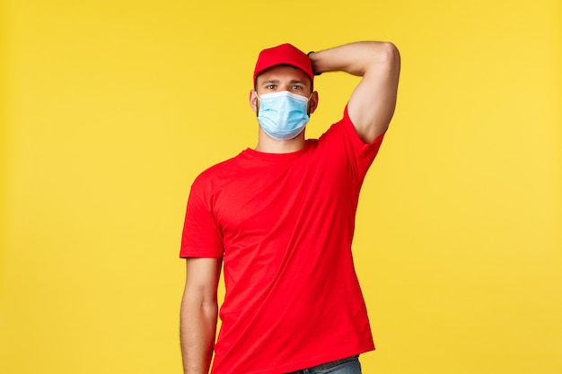 Express delivery during pandemic, covid-19, safe shipping, online shopping concept. Humble handsome courier in red uniform, medical mask, scratch head, standing yellow background