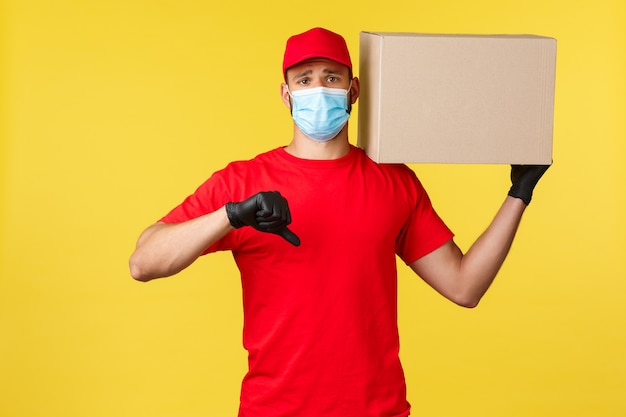 Express delivery during pandemic, covid-19, safe shipping, online shopping concept. Upset and disappointed courier in red uniform, medical mask, show thumb-down displeased, holding package