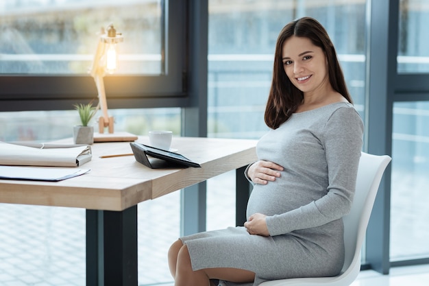 Expressing delight. Young pregnant woman sitting on chair while touching her stomach and smiling