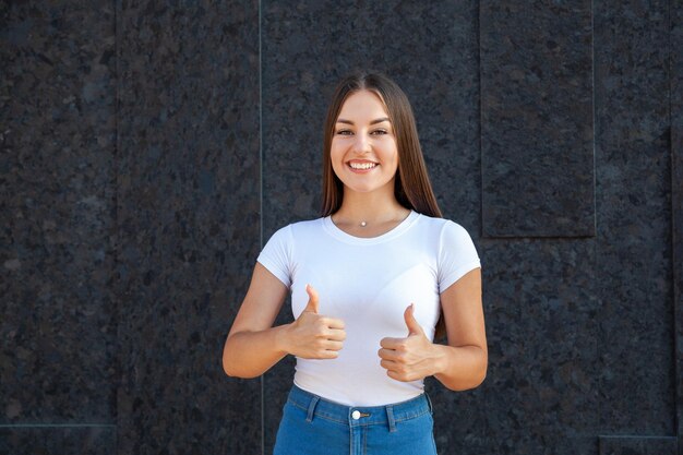 表現感情ジェスチャー人々の概念幸せな白人の女の子がカメラを見て親指を見せて、コピースペースで黒い背景に屋外で白いTシャツとジーンズに身を包んだ