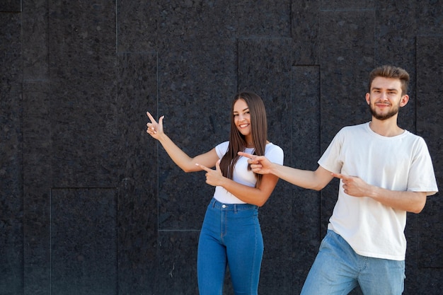 Expression emotions and people concept A happy Caucasian man and woman pointing with fingers to the left at the copy space and standing in white Tshirts and blue jeans on a black background