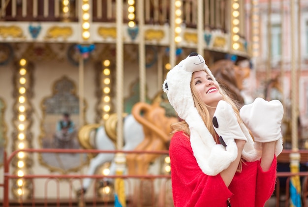 Expressive blonde woman wearing red knitted sweater and funny hat, posing at the background of carousel with lights
