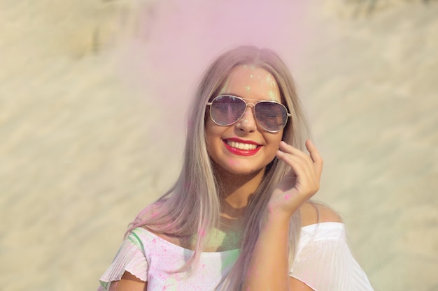 Выразительная блондинка в рубашке с обнаженными плечами и в очках играет с розовой сухой краской Холи в пустыне