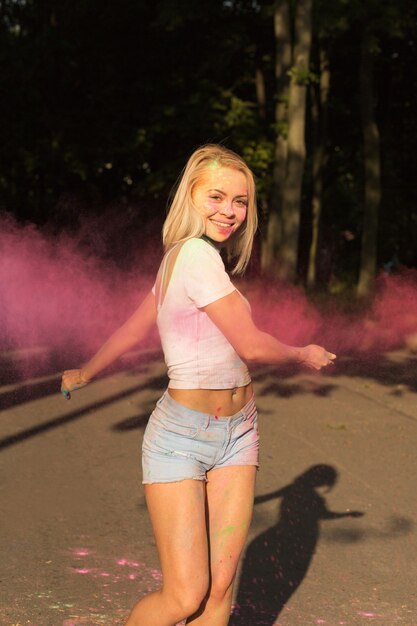 Expressive blonde woman in white t shirt and jeans shorts playing with pink Holi powder exploding around her