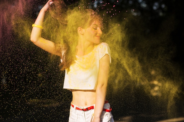 Expressive brunette woman posing in a cloud of yellow dry paint Holi at the park