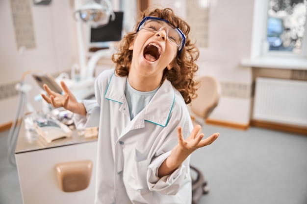 Expressive curly boy dressed as a doctor having his hands in the air while playing a mad scientist