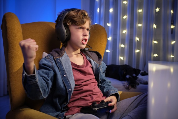 expressive curly haired boy in headphones sitting on comfortable armchair with gamepad while playing videogame and celebrating victory with raised arm and clenched fist near sleeping adorable miniatur