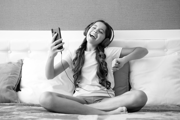 Expressive emotional excited teen girl Teenager child girl wearing headphones listening music on smart phone sitting on bed in her room