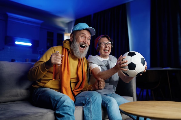 Expressive senior family couple watching football match on tv at home. Elderly husband and wife feeling overjoyed and excited screaming and cheering favorite soccer game