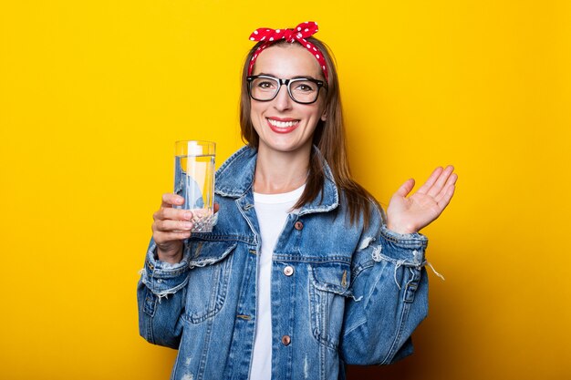 Giovane donna espressiva in jeans, occhiali e una fascia che tiene un bicchiere di acqua pulita