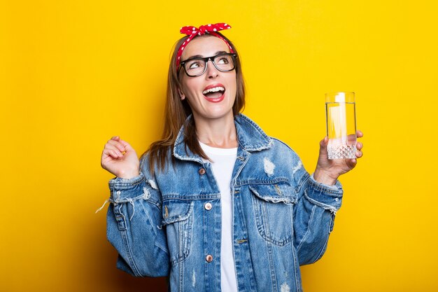 Expressive young woman in jeans, glasses and a headband holding a glass of clean water