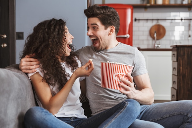 extatisch jong stel man en vrouw die thuis op de bank zitten en popcorn uit de emmer eten