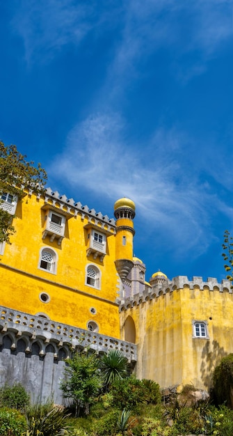 Exterior of the colorful yellow Pena Palace on fortified walls covered in vegetation and palm trees