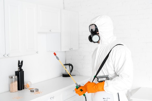 Exterminator in protective mask holding toxic equipment near kitchen cabinet