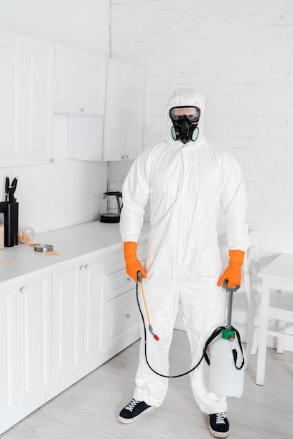 Exterminator in protective mask and uniform holding toxic equipment while standing near kitchen