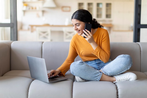 Externe zakelijke Indiase vrouw die telefonisch praat en thuis een laptop gebruikt