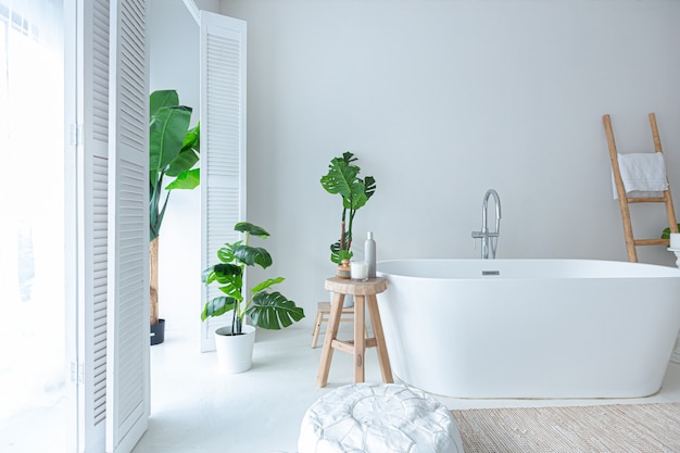 Очень белый и очень светлый минималистичный стильный элегантный интерьер ванной комнаты с современной ванной, зелеными растениями и деревянными элементами.