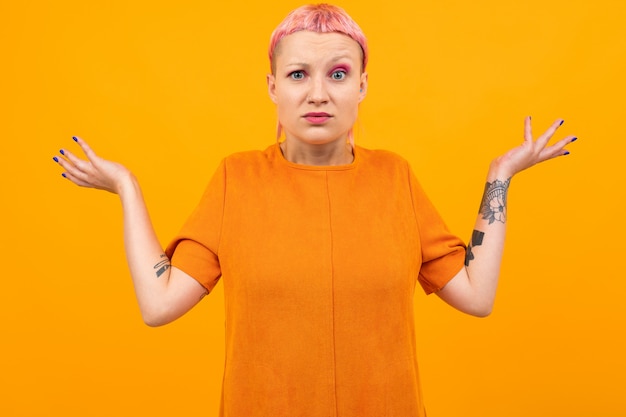 Extraordinary beautiful woman with short pink hair and big tattoo on her hand doesn't understand something isolated on orange