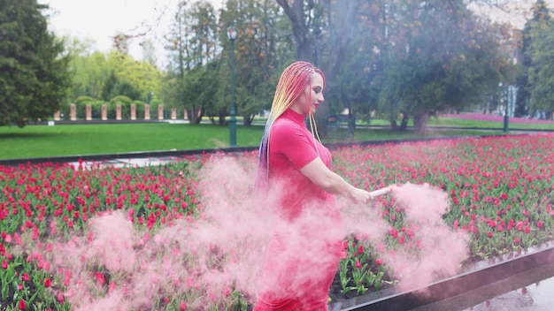 An extraordinary girl in a red dress with makeup and colored braids. Cute smiling and posing in thick pink smoke in the park in the rain