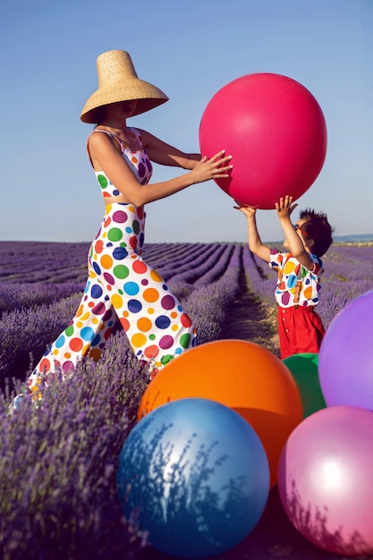 Photo extravagant woman mother in a white suit with multicolored polka dots ,and hat stands in a lavender field with her son in a multicolored clown costume