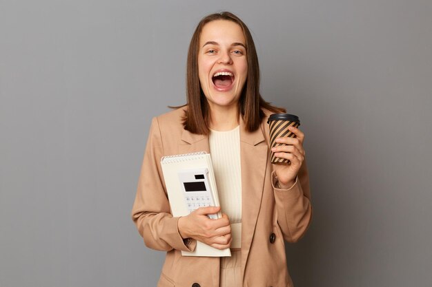 Extreem gelukkige blanke vrouw in beige jas met organizer en rekenmachine geïsoleerd over grijze achtergrond goed nieuws horend koffie drinken om haar succes te verheugen