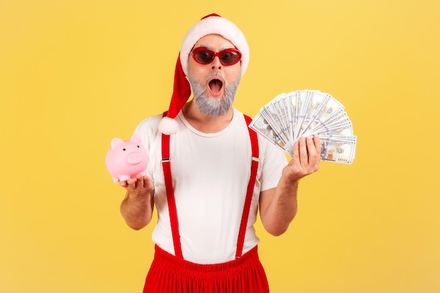 Extreem geschokt volwassen man in stijlvolle zonnebril en kerstman hoed met spaarvarken en dollar biljetten kijken camera aanbetaling cashback Indoor studio opname geïsoleerd op gele achtergrond