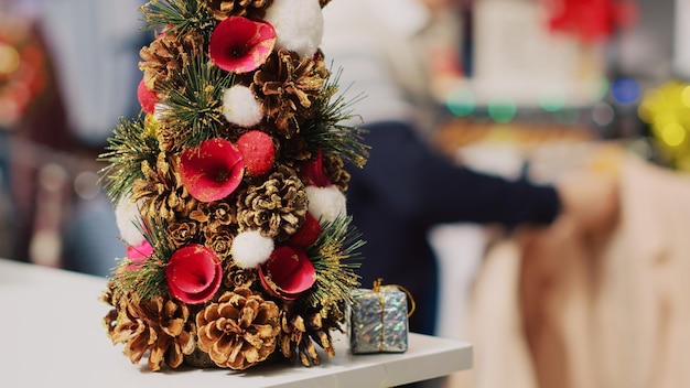 Extreme close-up shot van feestelijke miniatuur kerstboom versierd met dennenappels en bloemen zittend op de toonbank van de kledingwinkel tijdens de wintervakantie. Kerstdecor in modeboetiek