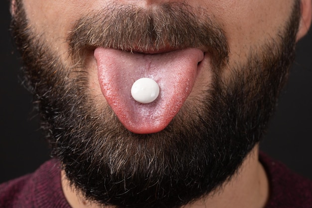 Extreme close-up van witte ronde pil op tong van bebaarde man gezicht met donkere snor op zwarte achtergrond nemen vitamines pijnstiller