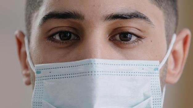 Extreme close up young man surgeon hispanic male face in medical mask with dark eyes looking at