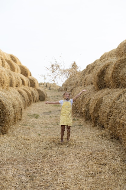 Extremely excited female kid throwing hay up standing close to\
heaps of haystacks on field looking up wearing sundress having fun\
away from city on field full of golden hay