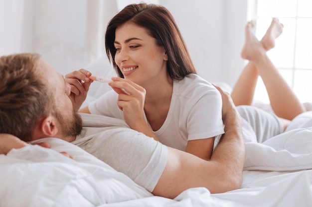 妊娠中のミレニアル世代のブルネットが夫と一緒にベッドに横たわっていて、妊娠検査陽性であるという事実に非常に満足しています