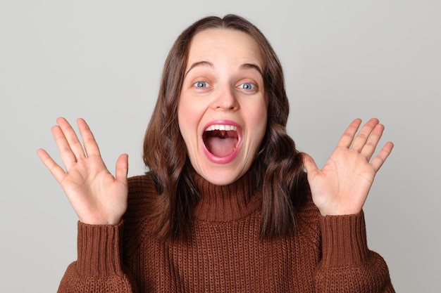 Extremely happy Caucasian woman wearing brown sweater standing isolated over light gray background raising her arms screaming with positive emotions rejoicing