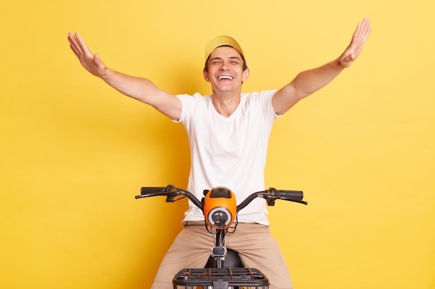 매우 행복한 백인 청년은 흰색 티셔츠와 모자를 쓰고 전기 자전거를 타고 노란색 배경에서 격리된 여가 시간을 즐기고 있습니다.