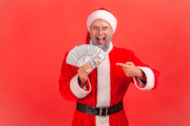 Extremely happy excited man in santa claus costume pointing finger at dollar banknotes he holding in hand looking at camera with big eyes and open mouth indoor studio shot isolated on red background