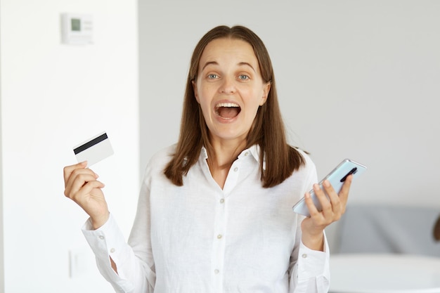 Extremely happy excited woman wearing white shirt standing indoor in light room with smart phone and credit card in hands, winning lottery, online shopping or banking.