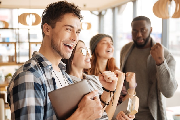 Extremely happy. Handsome youthful man is smiling and expressing happiness with his joyful fellows on background.